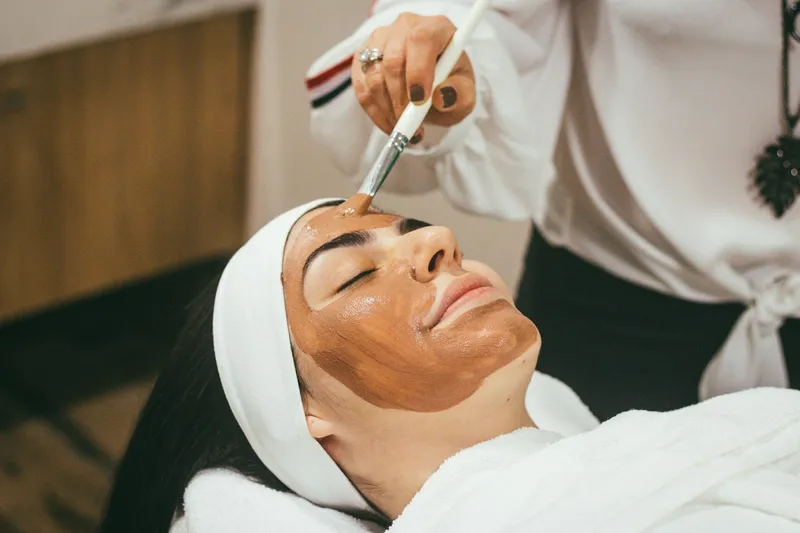 Cosmetóloga aplica con brocha una mascarilla oscura en el rostro de una clienta recostada